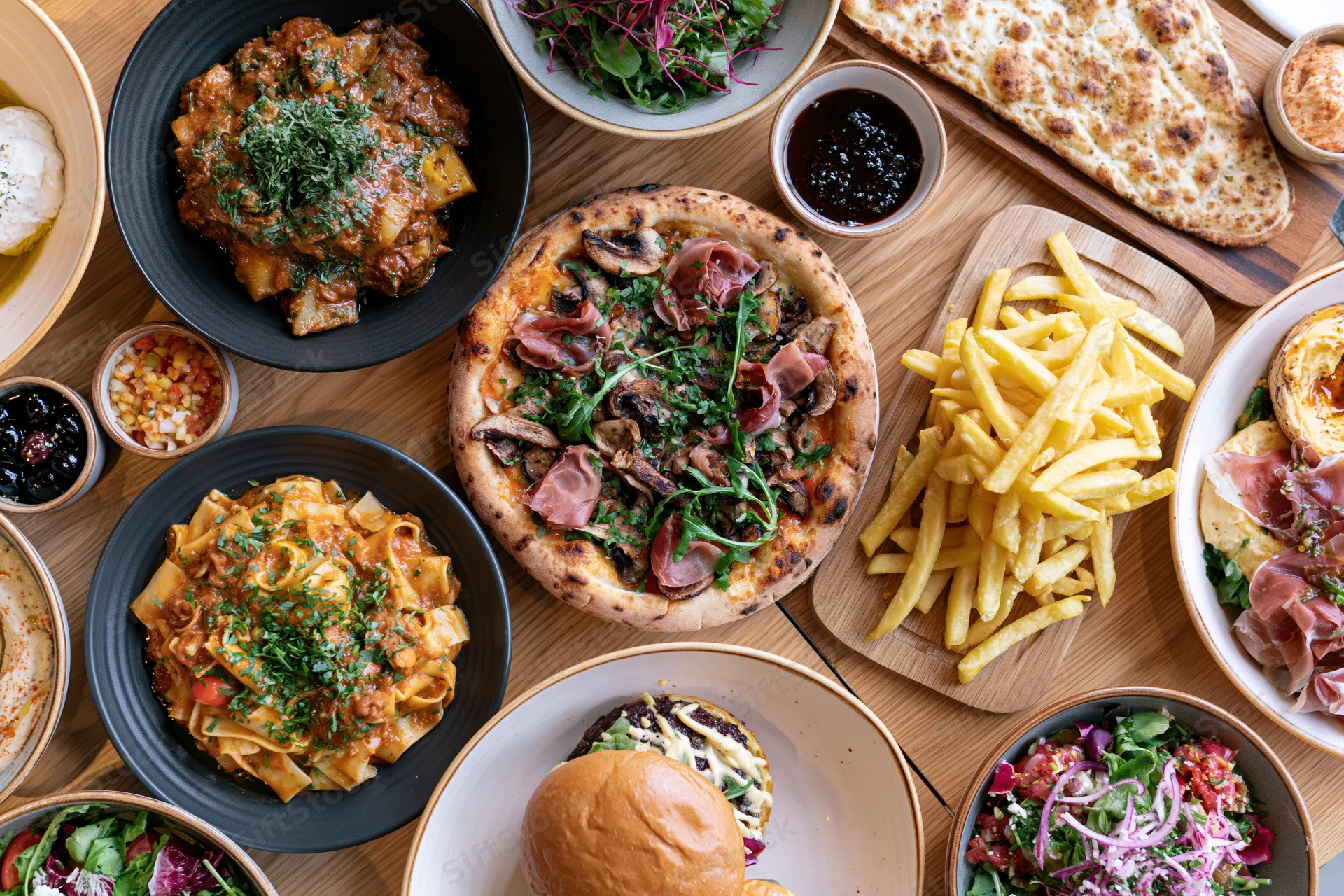 Overhead view of a restaurant table filled with various dishes including pizza, pasta, burger, fries, salad, and naan bread arranged for dining
