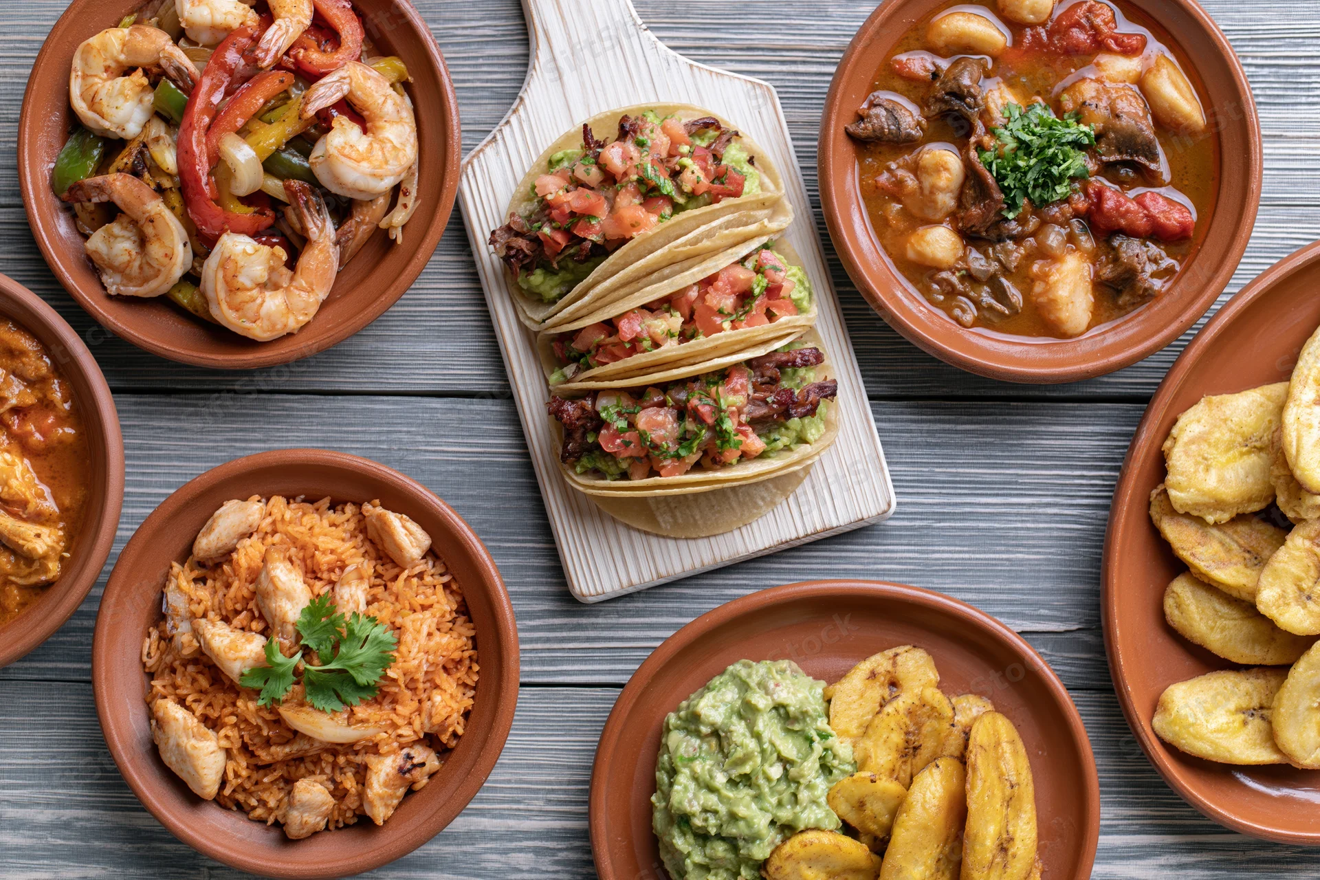 Overhead flat-lay spread of traditional mexican dishes on wooden table including tacos, shrimp, rice, beans, guacamole, and plantains