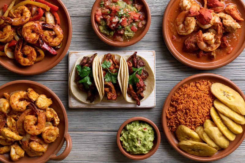 Overhead flat-lay of traditional mexican dishes including tacos, shrimp, rice, guacamole, and plantains on a wooden table