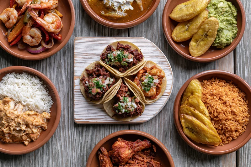 Overhead flat-lay of traditional mexican dishes arranged on wooden table with tacos, rice, beans, shrimp, chicken, guacamole, and plantains
