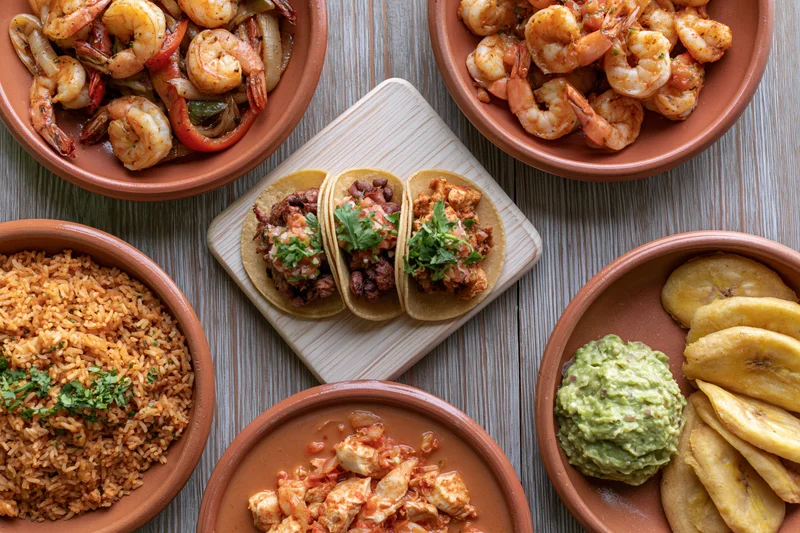 Overhead flat-lay of traditional mexican dishes including tacos, shrimp, rice, guacamole and plantains on a wooden table