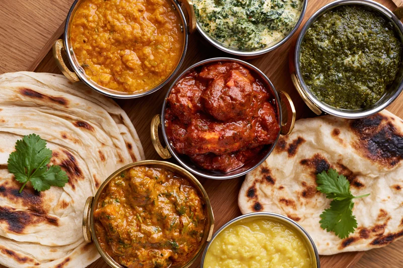 Overhead flat lay of indian curry spread with multiple brass bowls and naan bread on wooden tray