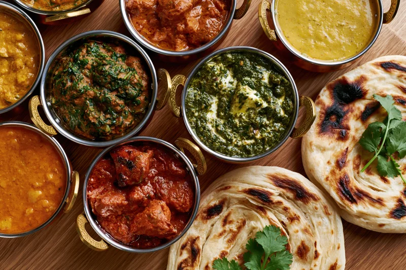Overhead flat lay of indian curry dishes in brass bowls with naan bread on wooden surface