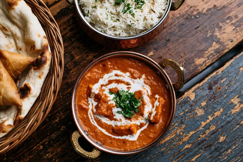 Butter chicken curry with cream swirl and cilantro garnish served in copper pot alongside basmati rice and naan bread in woven basket on rustic wooden table