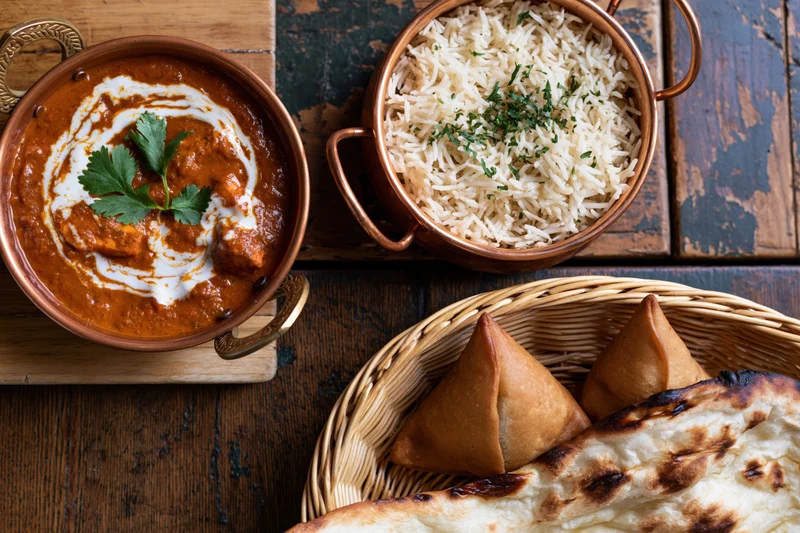 Indian meal with curry, rice, naan and samosas arranged on wooden table in overhead flat lay