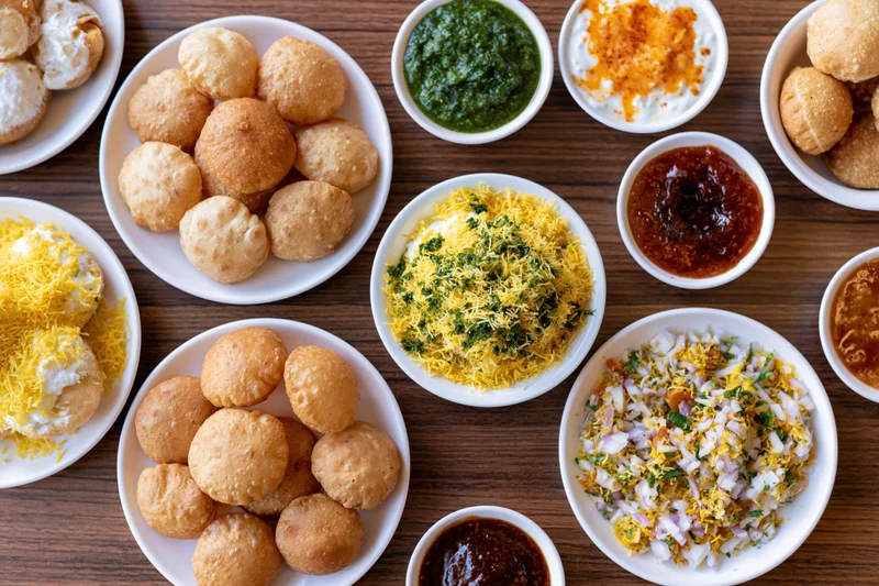 Overhead flat lay of pani puri and dahi street food arranged on wooden table with chutneys and toppings in white bowls