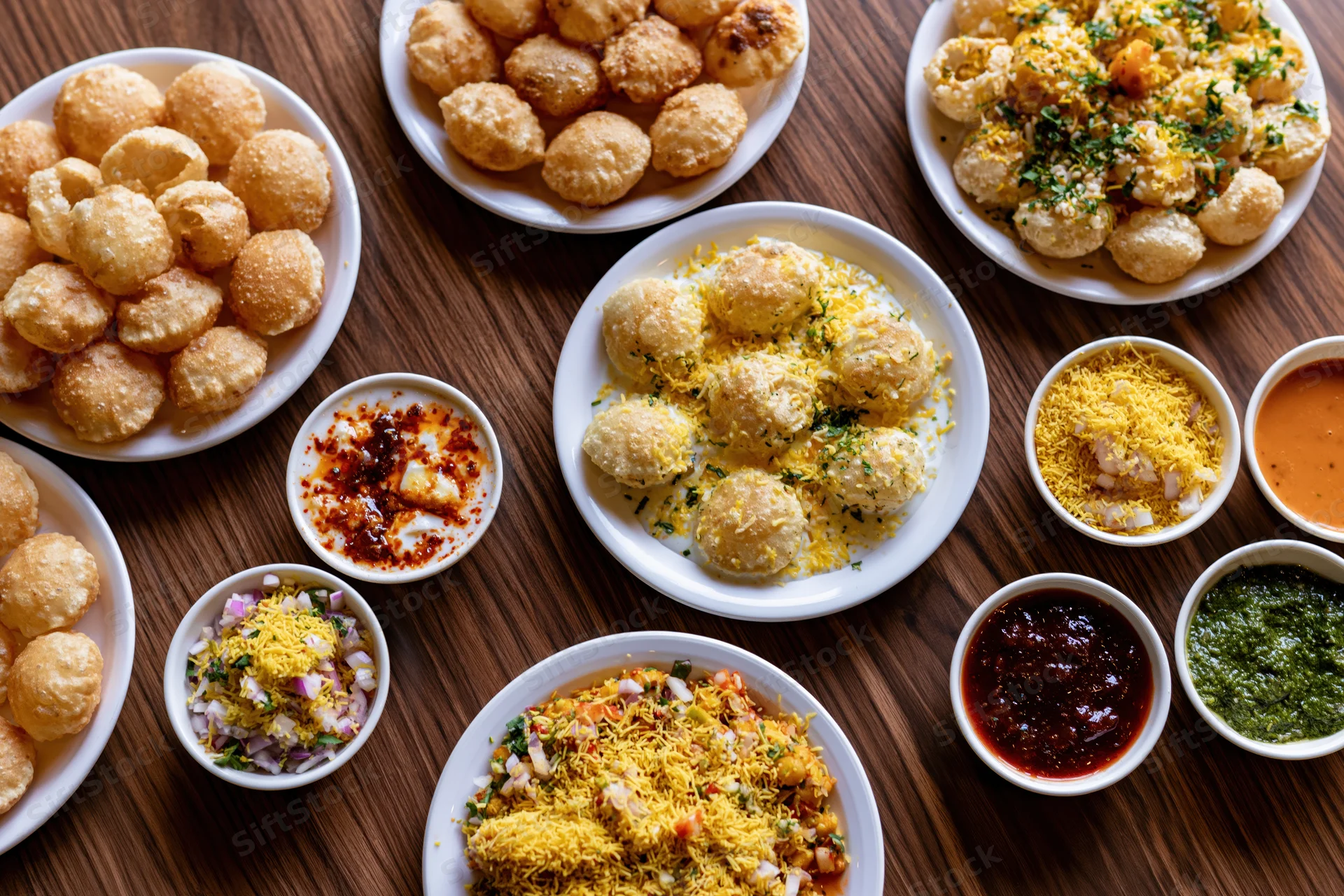 Overhead flat lay of indian street food pani puri and dahi puri served on white plates with chutneys and sev on wooden table