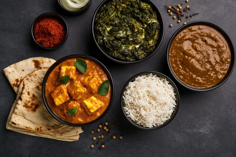 Overhead flat lay of indian vegetarian meal with paneer curry, palak, dal, rice, naan, and spices on dark surface