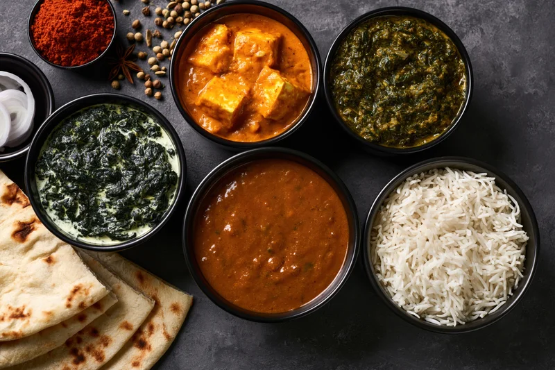 Overhead flat lay of indian vegetarian meal with curry, rice, naan and spices in black bowls on dark surface