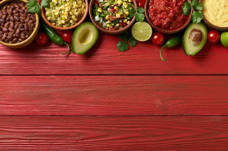 Mexican dips and condiments arranged in bowls with avocado, lime, and herbs on a red wooden table, overhead flat lay with copyspace below