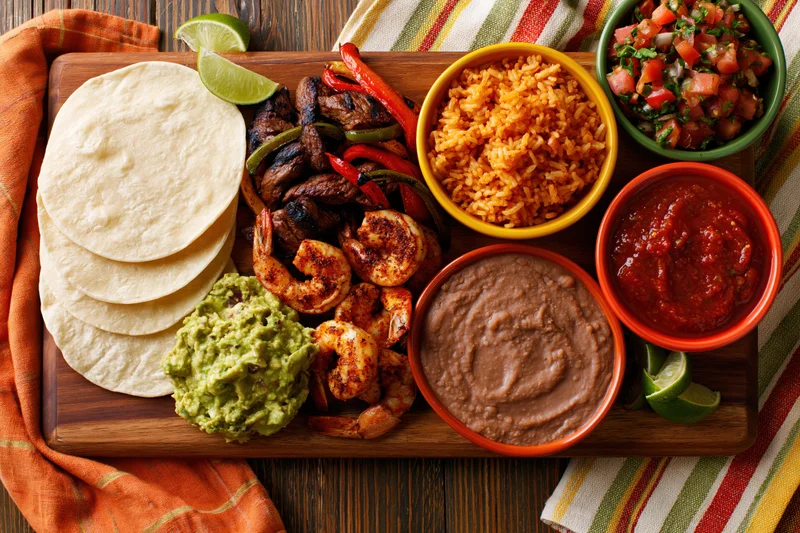 Overhead flat lay of mexican fajita spread with tortillas, shrimp, steak, guacamole, refried beans, rice, salsa and pico de gallo on wooden cutting board