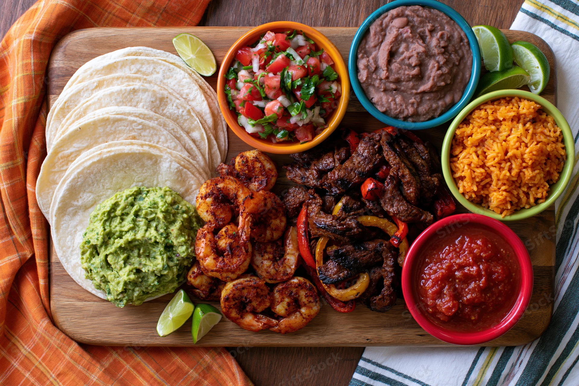 Overhead flat lay of mexican fajita spread with tortillas, guacamole, shrimp, steak, rice, beans, salsa and lime on wooden cutting board