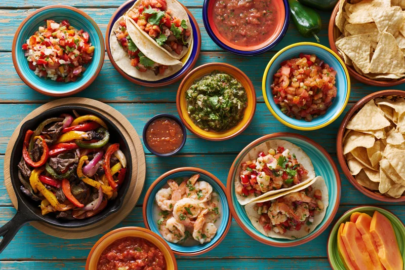 Overhead flat lay of mexican food spread on turquoise wooden table with tacos, salsa, guacamole, chips, fajitas and shrimp