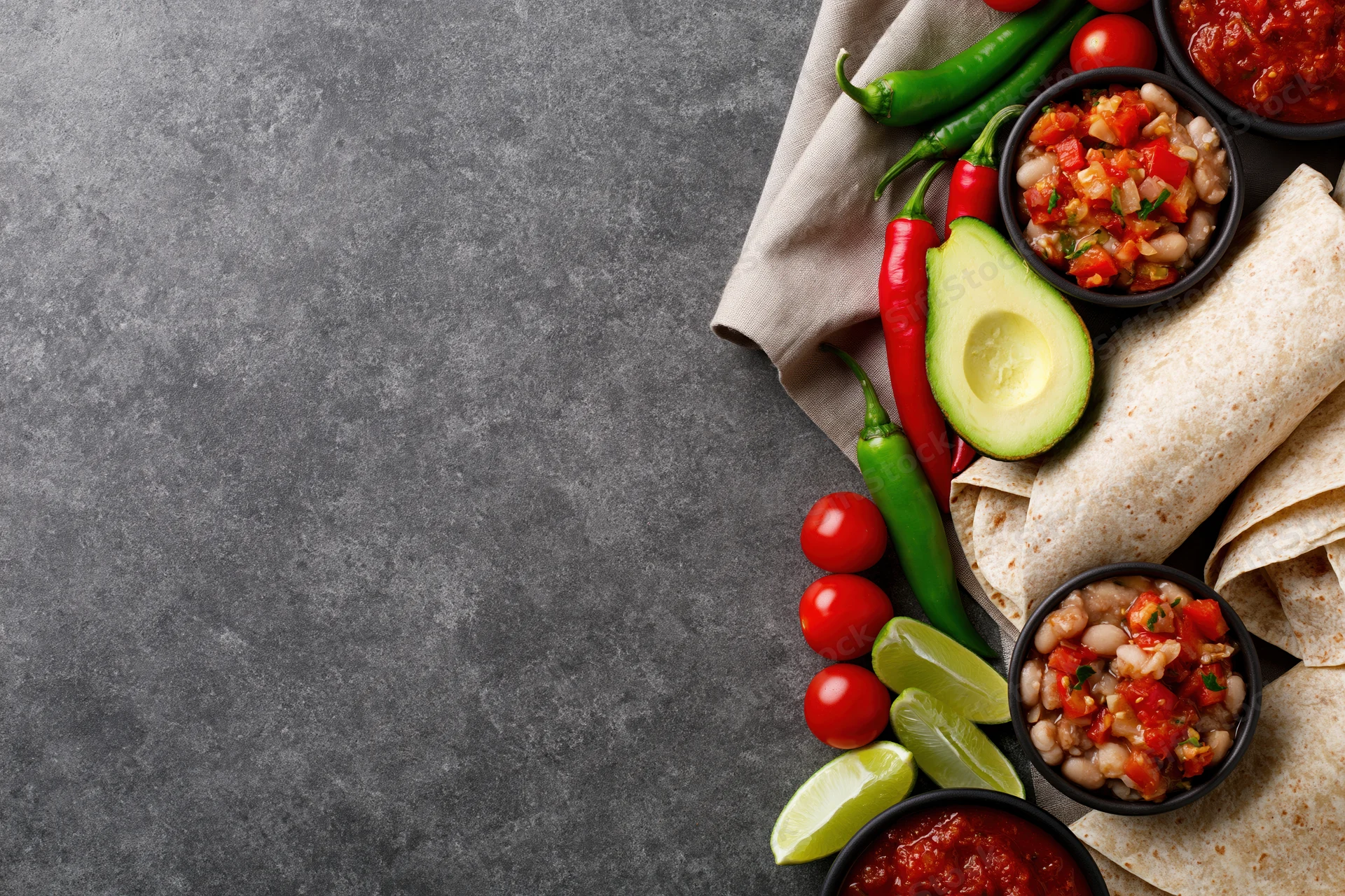 Mexican food spread arranged along the right side of a gray concrete surface with tortillas, avocado, chili peppers, tomatoes, lime, beans, and salsa in bowls, creating a border with copyspace on the left