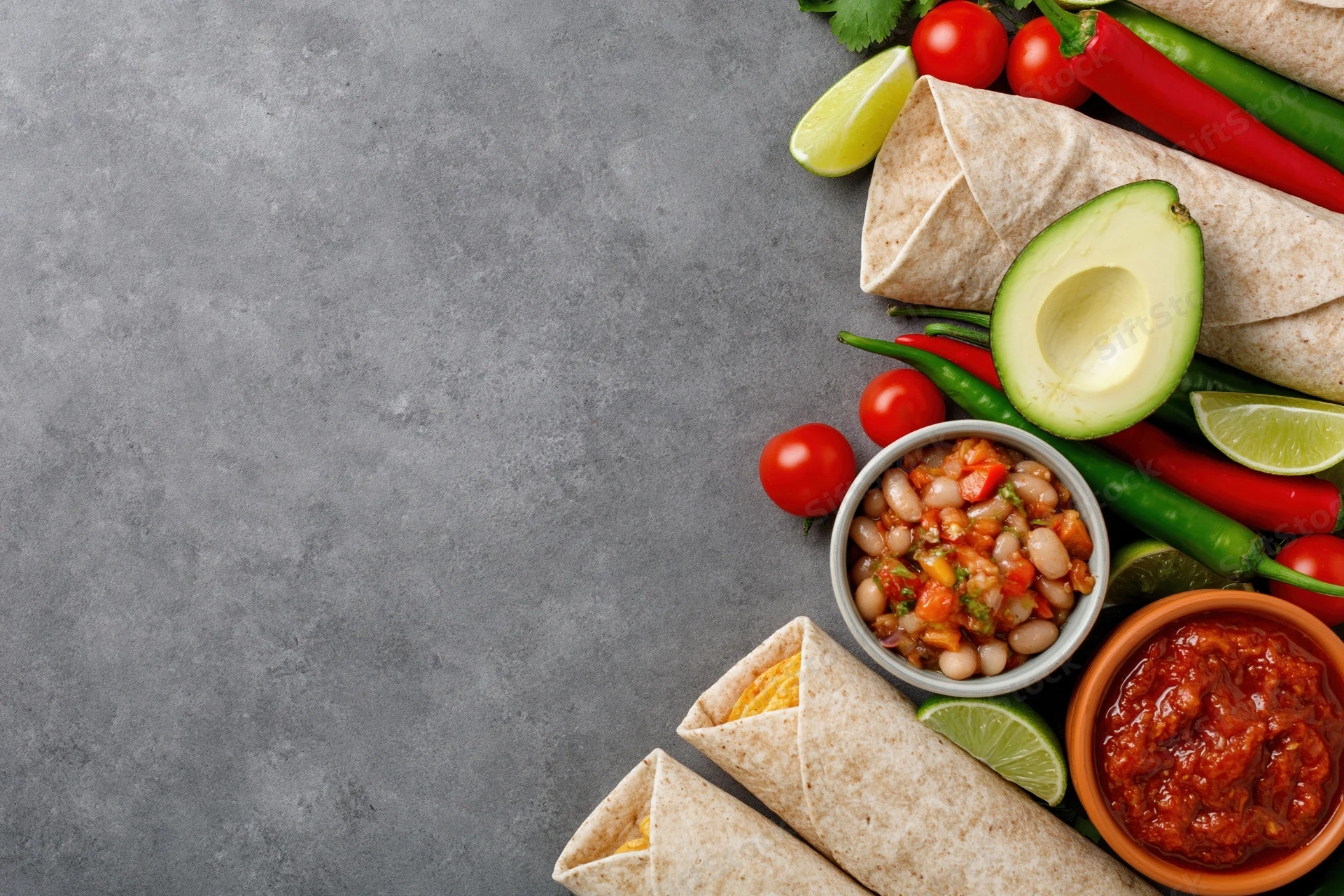 Mexican food spread with burritos, avocado, salsa, beans, lime, and peppers arranged along the right side of a gray surface