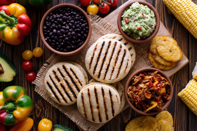 Grilled venezuelan arepas with black beans, guacamole, pulled pork, and plantains arranged on a wooden table with vegetables and corn in a flat lay overhead view