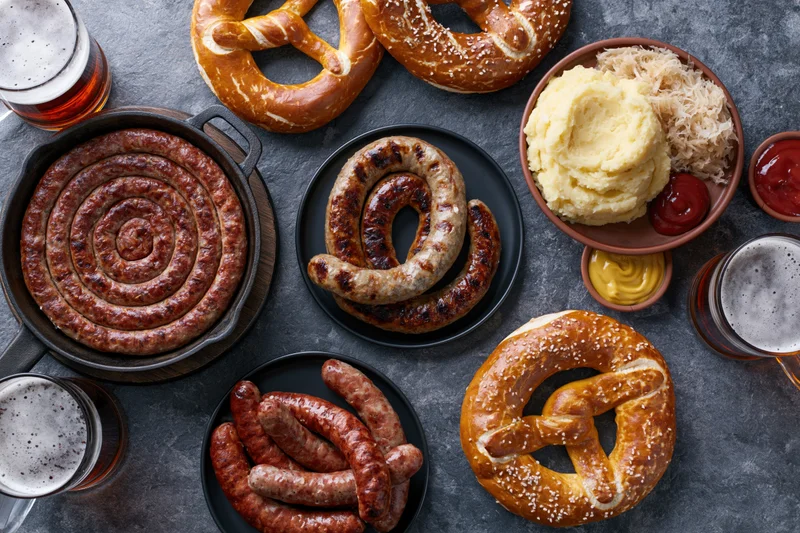Overhead flat lay of german oktoberfest food spread with sausages, pretzels, beer, and condiments on dark stone surface