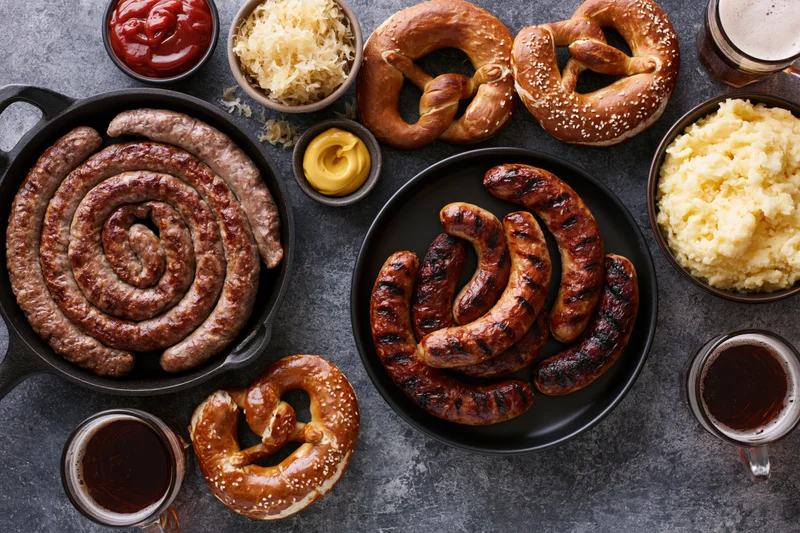 Grilled sausages and pretzels with beer and sides arranged on dark stone surface for Oktoberfest celebration