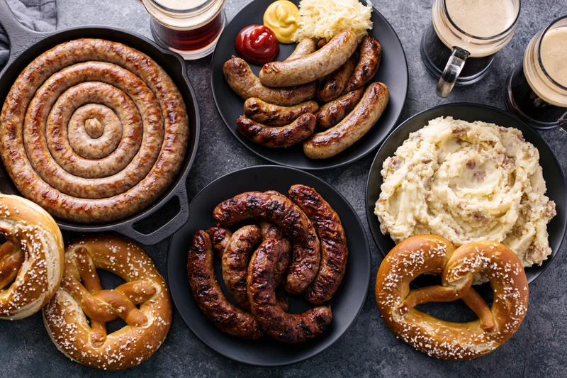 Overhead flat lay of german oktoberfest food spread with sausages, pretzels, mashed potato, and beer on dark surface