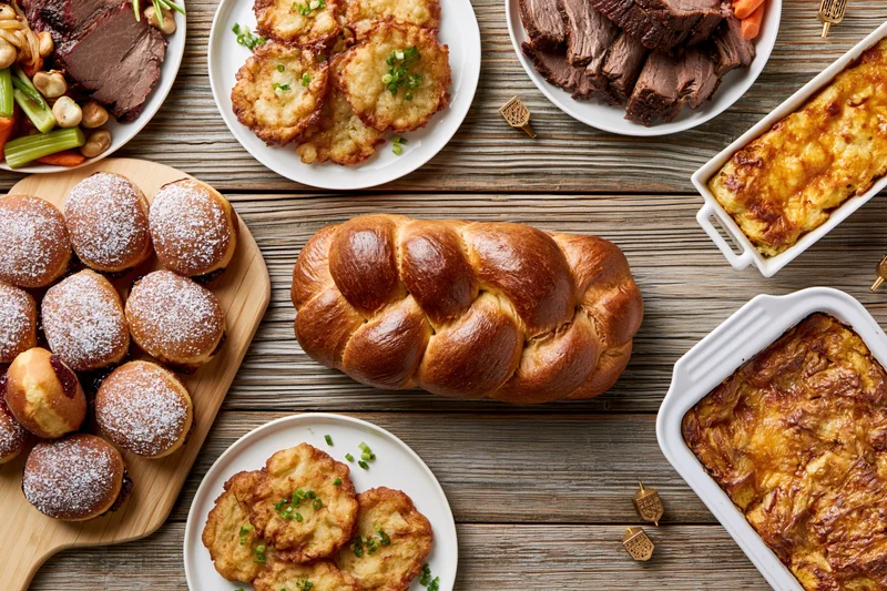 Overhead view of a hanukkah holiday spread with challah bread, latkes, sufganiyot, roast beef, vegetables, and casseroles on a rustic wooden table