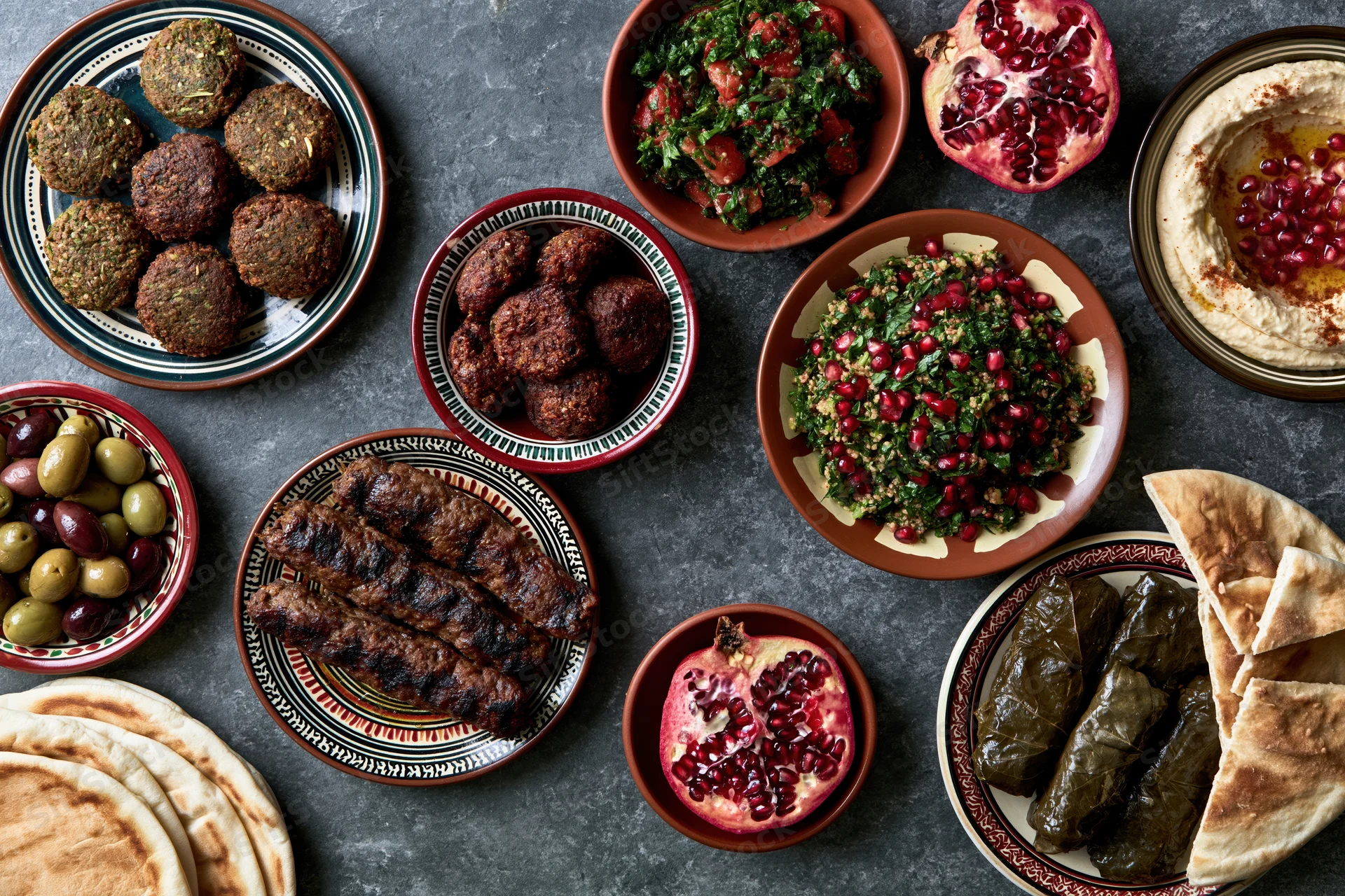Overhead flat lay of a lebanese mezze spread with hummus, tabbouleh, falafel, kibbeh, olives, pomegranate, dolma, and pita on a dark surface