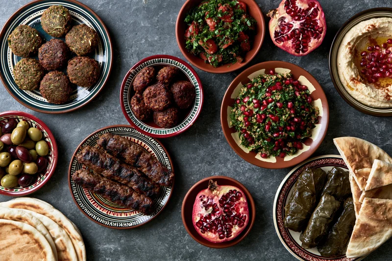 Overhead flat lay of a lebanese mezze spread with hummus, tabbouleh, falafel, kibbeh, olives, pomegranate, dolma, and pita on a dark surface