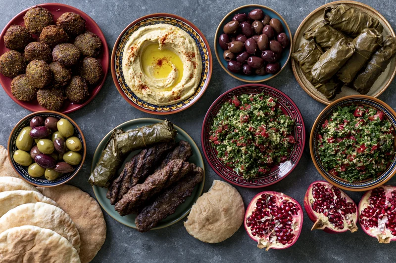 Overhead flat lay of a lebanese mezze spread with hummus, falafel, tabbouleh, olives, pomegranate, dolma, kibbeh, pita and meat on a dark stone surface