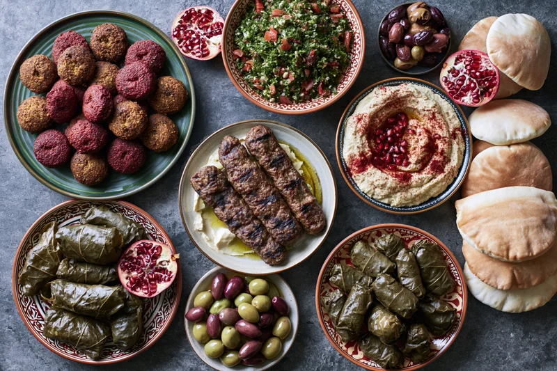 Overhead flat lay of a lebanese mezze spread with hummus, falafel, kofta, tabbouleh, dolma, pita, olives and pomegranate on a dark stone table