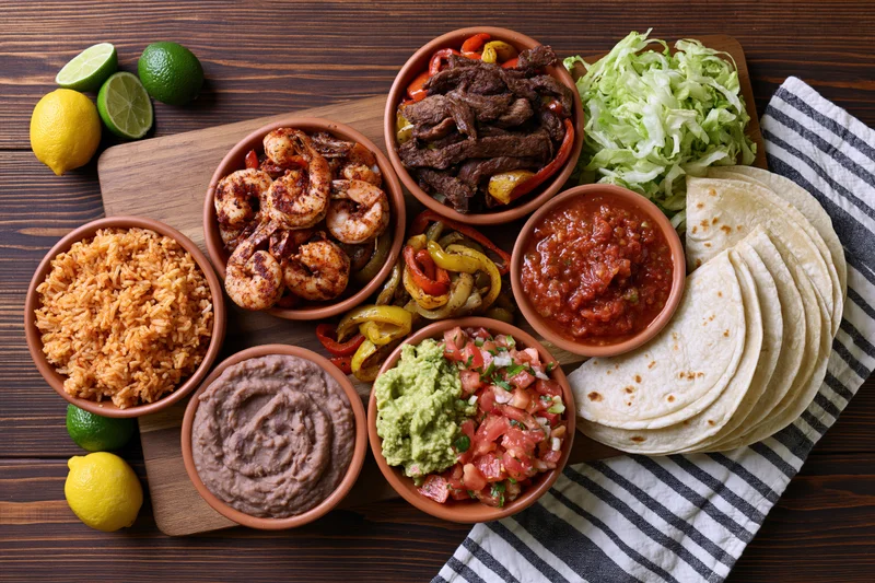 Overhead flat lay of a mexican fajita spread with tortillas, rice, beans, guacamole, salsa, and vegetables on a wooden table