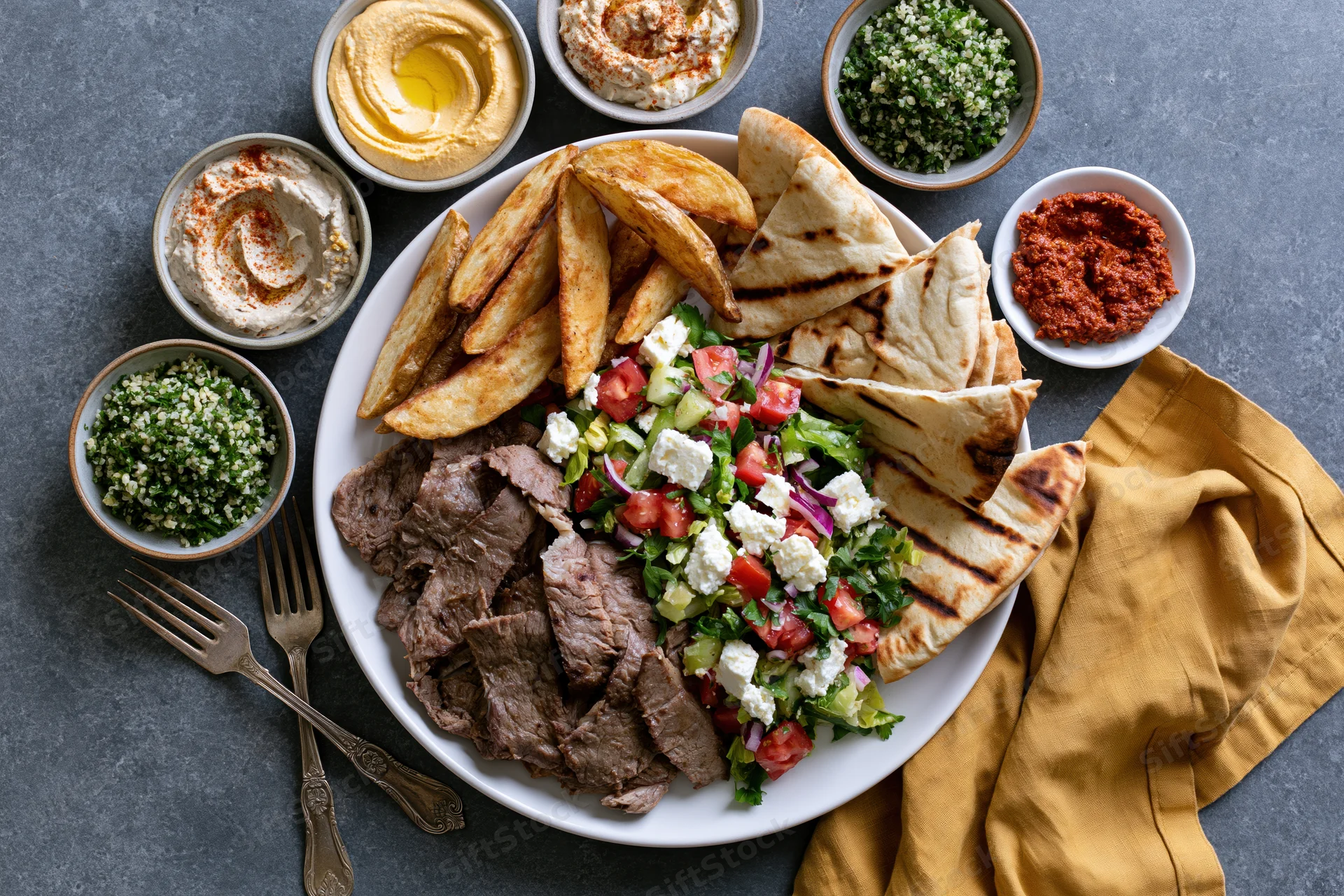 Overhead flat lay of a middle eastern mezze platter with grilled pita, sliced meat, salad, and assorted dips on a dark surface