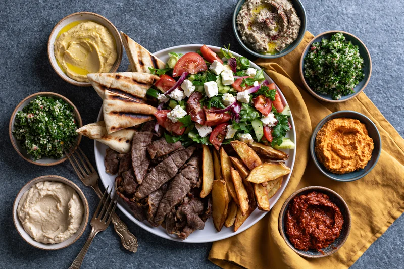 Overhead flat lay of a middle eastern mezze spread with grilled meat, salad, pita, fries, and various dips on a dark stone table