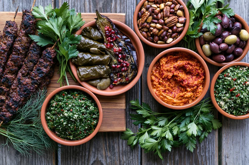 Overhead flat lay of a middle eastern mezze spread with grilled kebabs, dolma, hummus, tabbouleh, olives and nuts on a wooden board