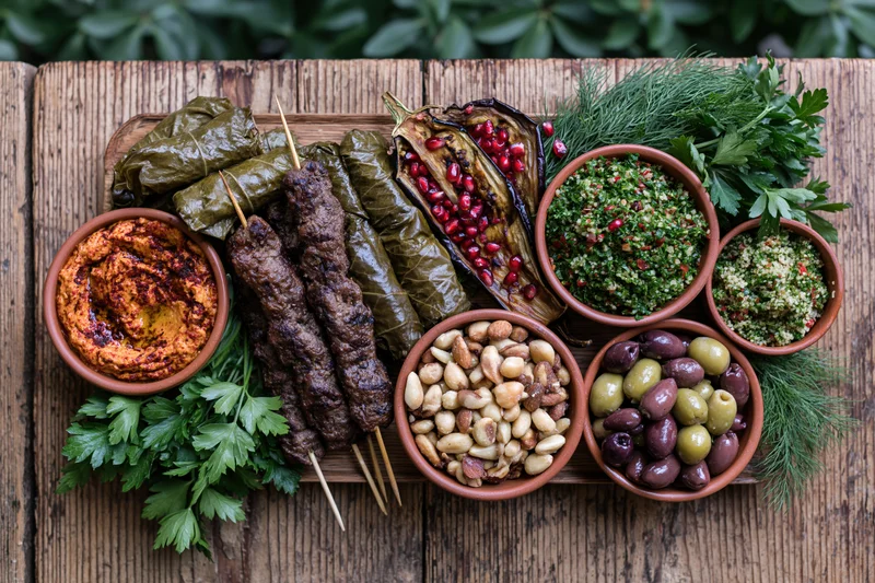 Overhead flat lay of a middle eastern mezze spread on a wooden table with grilled kebabs, dolma, hummus, olives, nuts, and fresh herbs arranged around bowls of dips and garnishes