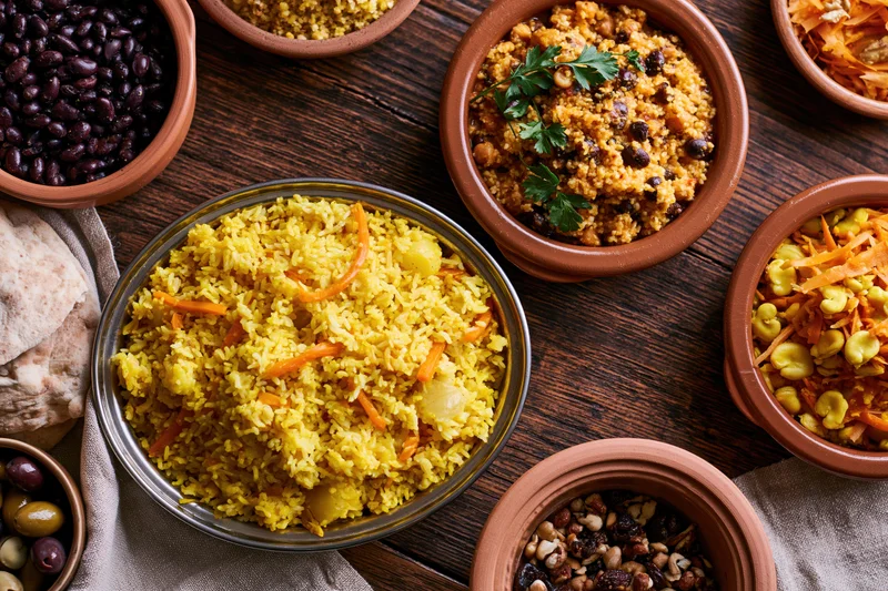 Overhead flat lay of a middle eastern and north african meal spread with rice, couscous, beans, vegetables and bread on a wooden table