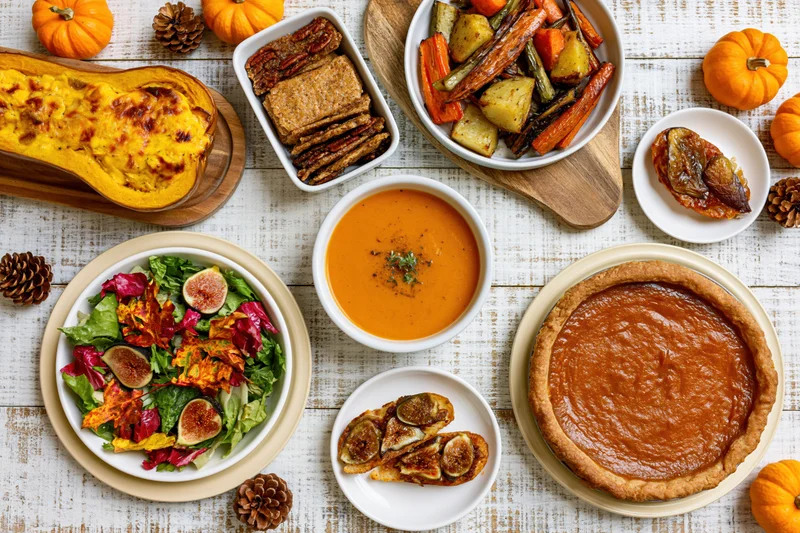 Overhead flat lay of thanksgiving dishes including pumpkin pie, salad, soup, roasted vegetables, and garnishes on a whitewashed wooden table