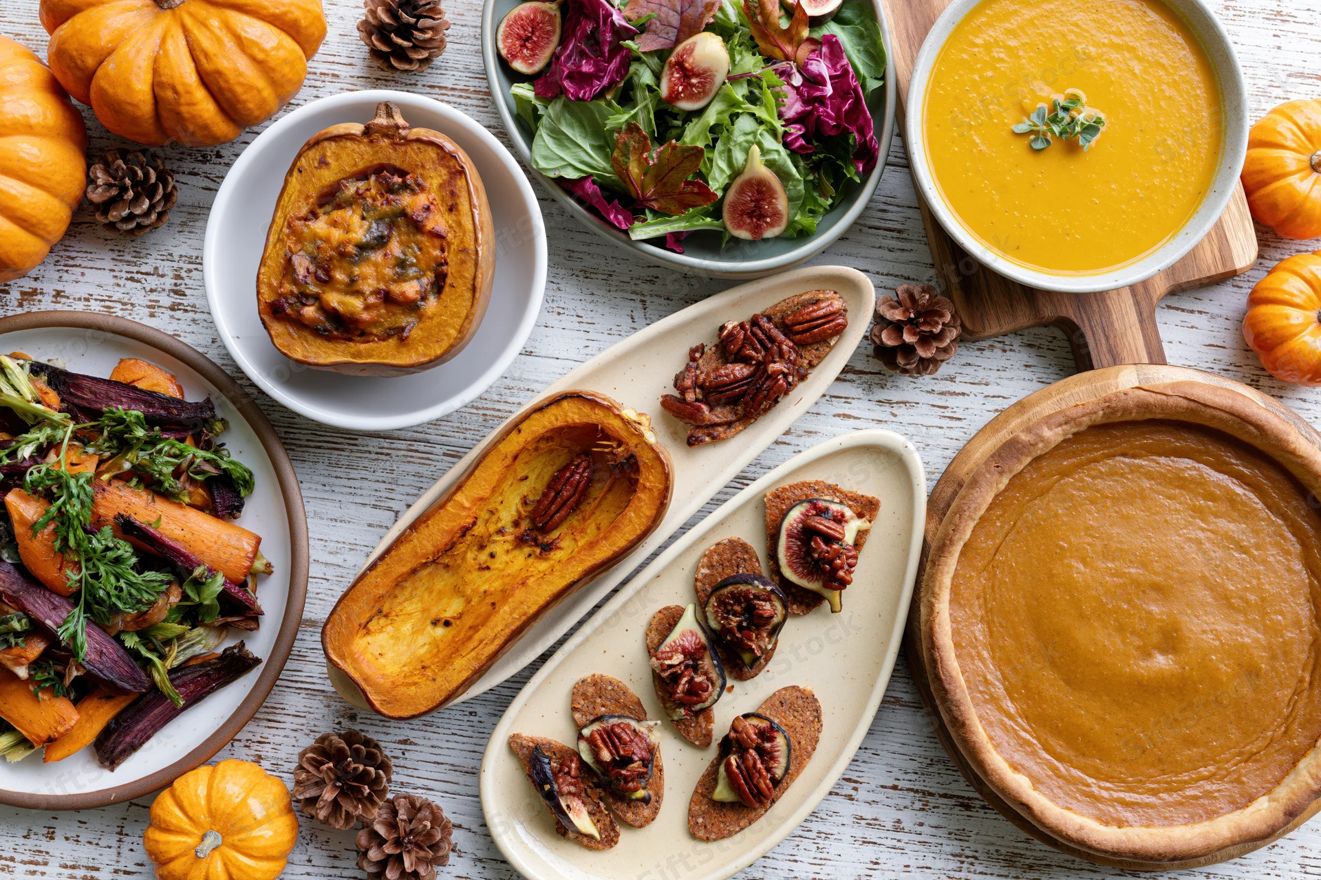 Overhead flat lay of thanksgiving dishes including pumpkin pie, soup, salad, and roasted vegetables on a whitewashed wooden table
