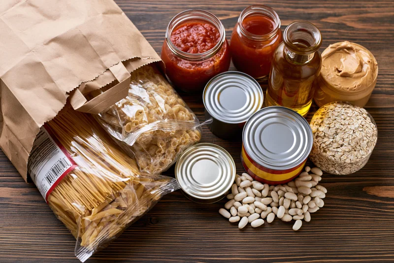 Overhead flat lay of groceries including pasta, beans, canned goods, jars and oats arranged on a dark wooden table