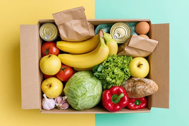 Open cardboard box filled with fresh fruits vegetables bread eggs and canned goods on yellow and teal background