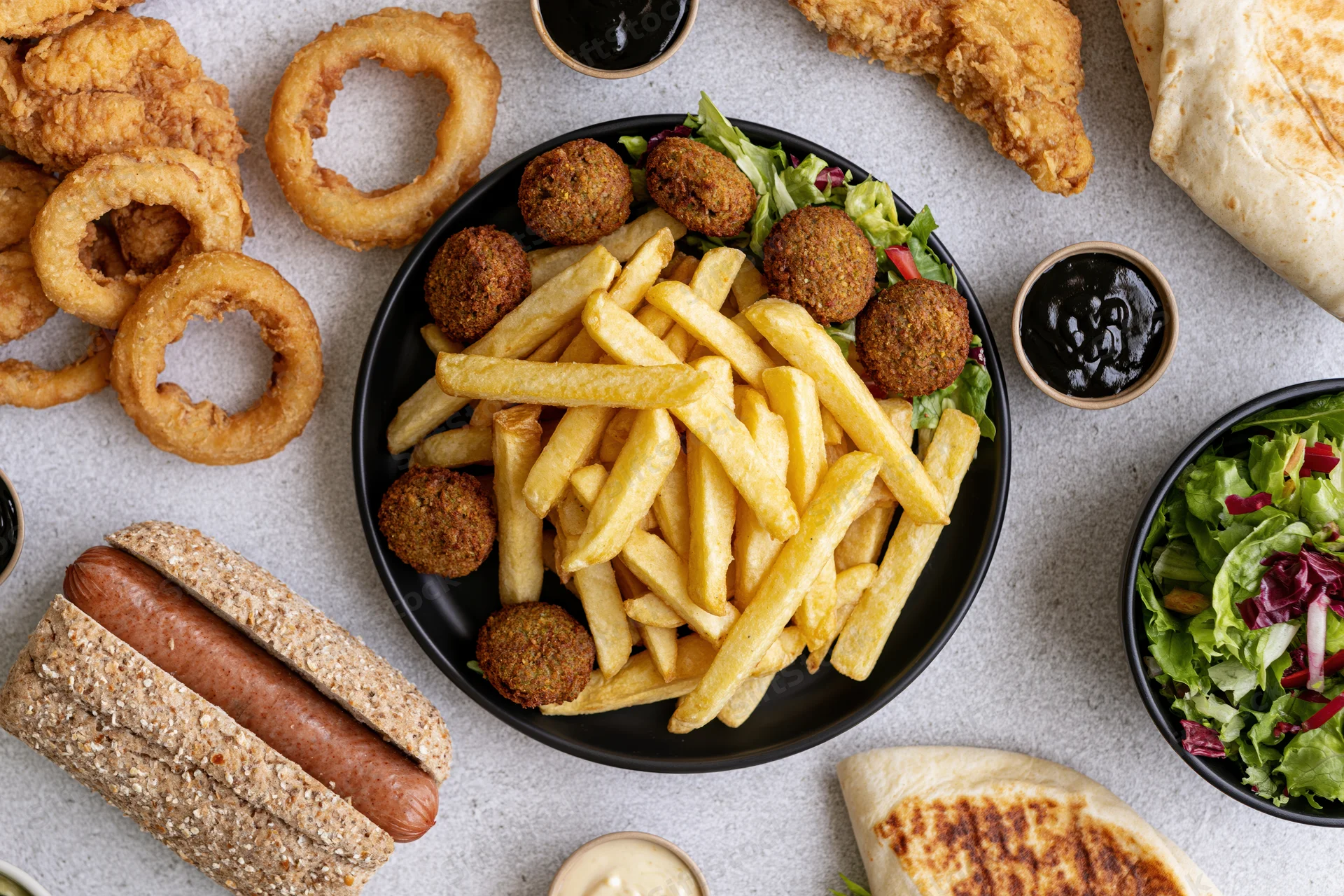 Overhead flat lay of a casual fast-food spread with fries, onion rings, hot dog, wrap, salad, and meatballs on a light gray surface
