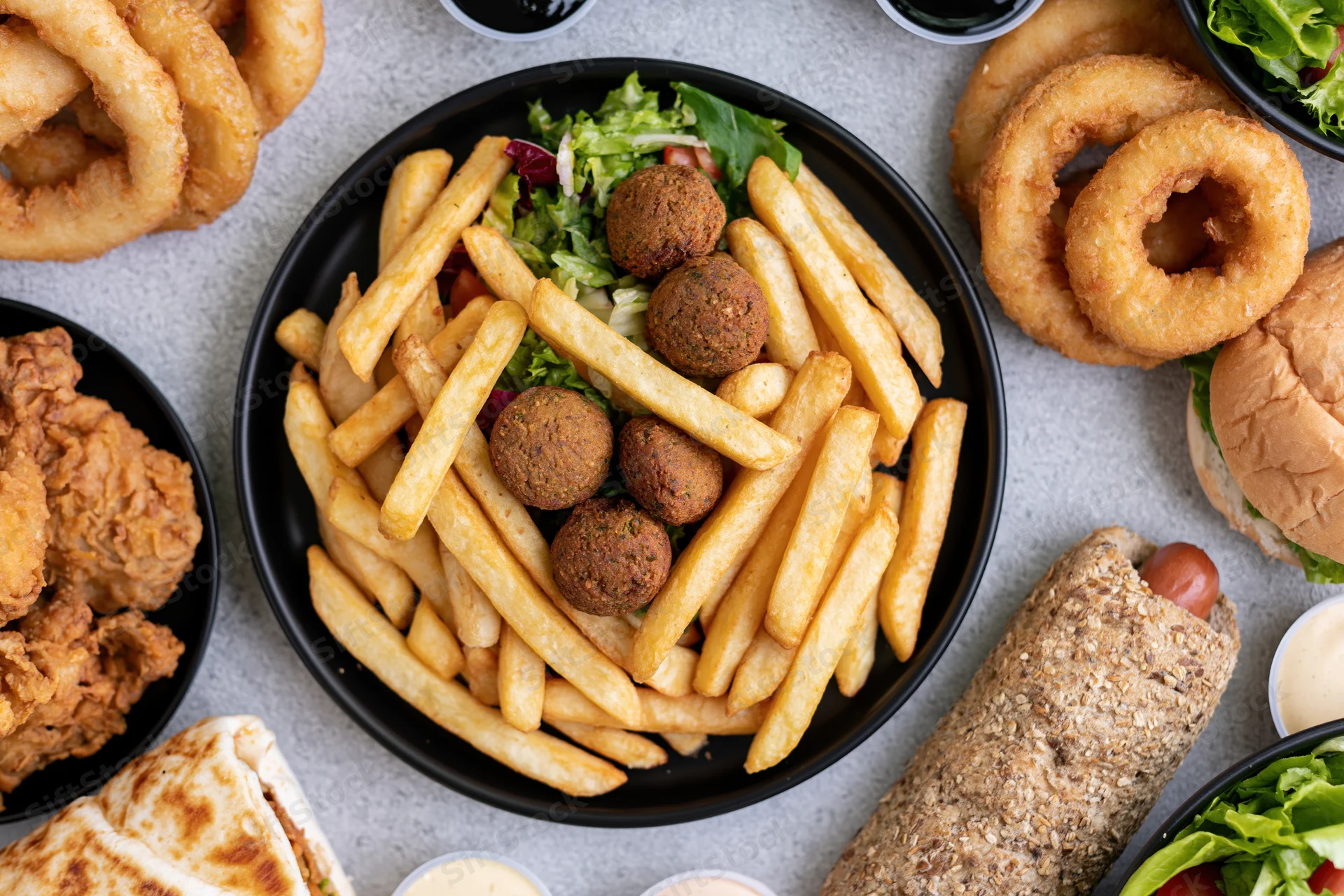 Overhead flat lay of a casual fast-food spread with fries, burgers, onion rings, chicken, falafel, and sauces on a gray surface