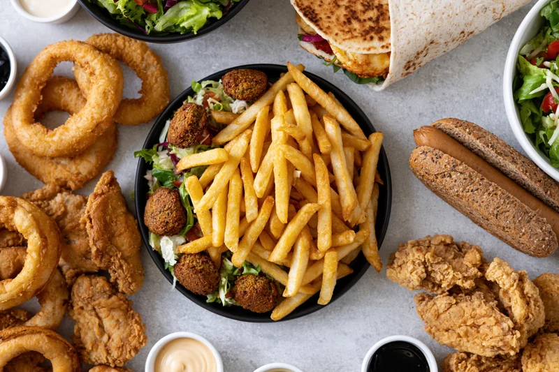 Overhead flat lay of fast food items including fries, onion rings, chicken, burger, wrap, and salad on a gray surface