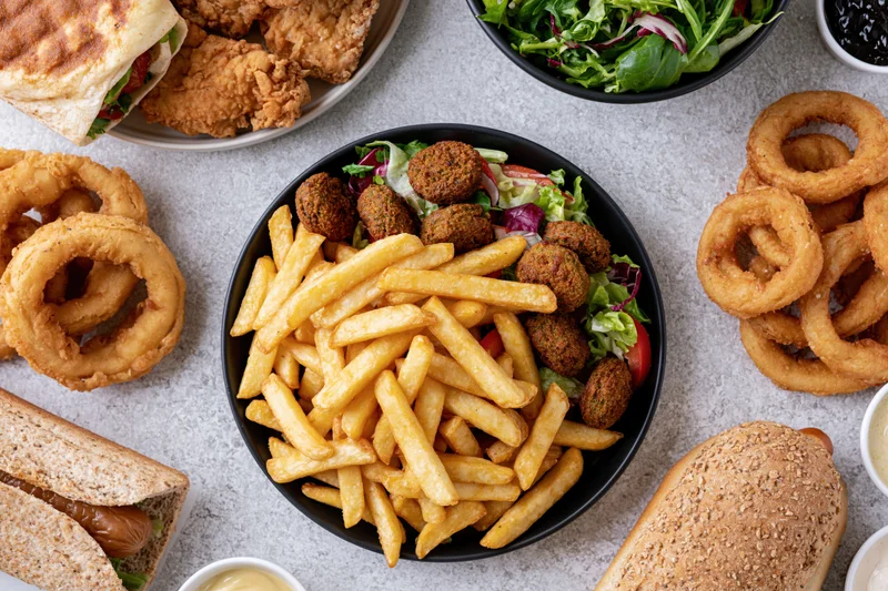 Overhead flat lay of a casual fast-food spread with fries, onion rings, burgers, sandwiches, and salads on a light gray textured surface