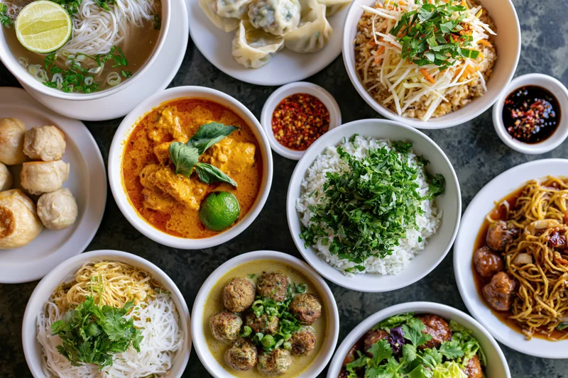 Overhead view of multiple asian dishes arranged on a dark table with bowls of rice, noodles, curry, dumplings and garnishes