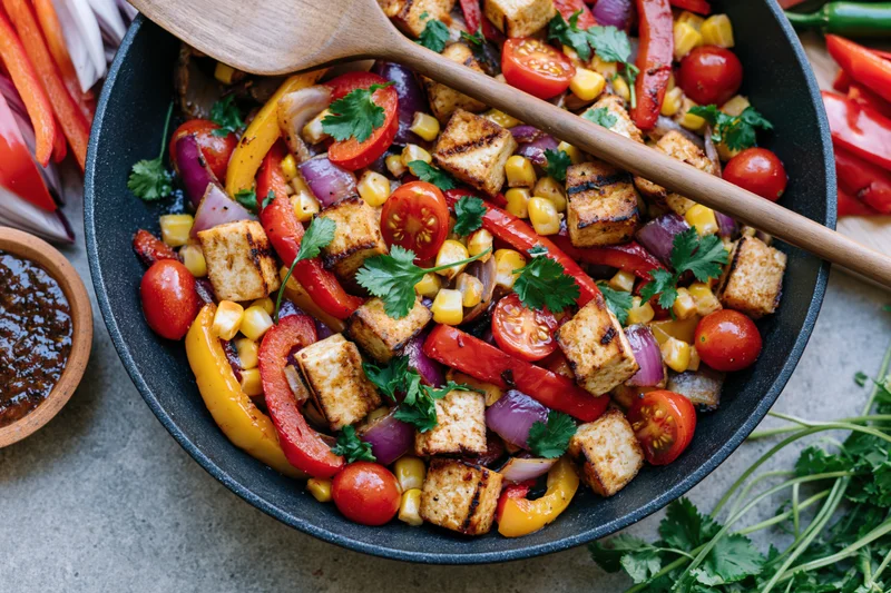 Overhead view of a dark skillet filled with sautéed tofu and colorful vegetables, garnished with cilantro and a wooden spoon resting on top