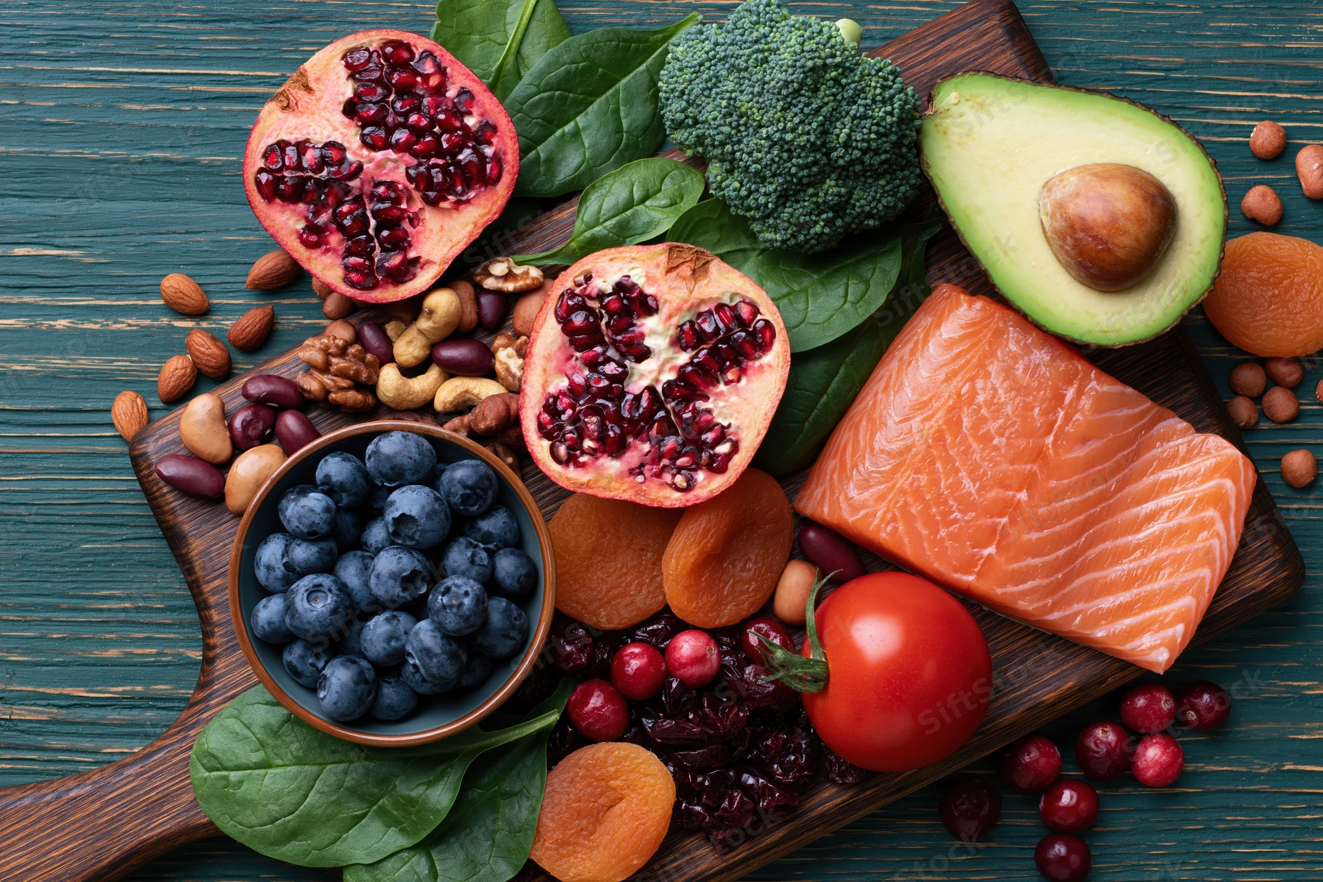 Salmon and avocado with pomegranate, blueberries, broccoli, and nuts arranged on a dark wooden cutting board in overhead flat lay