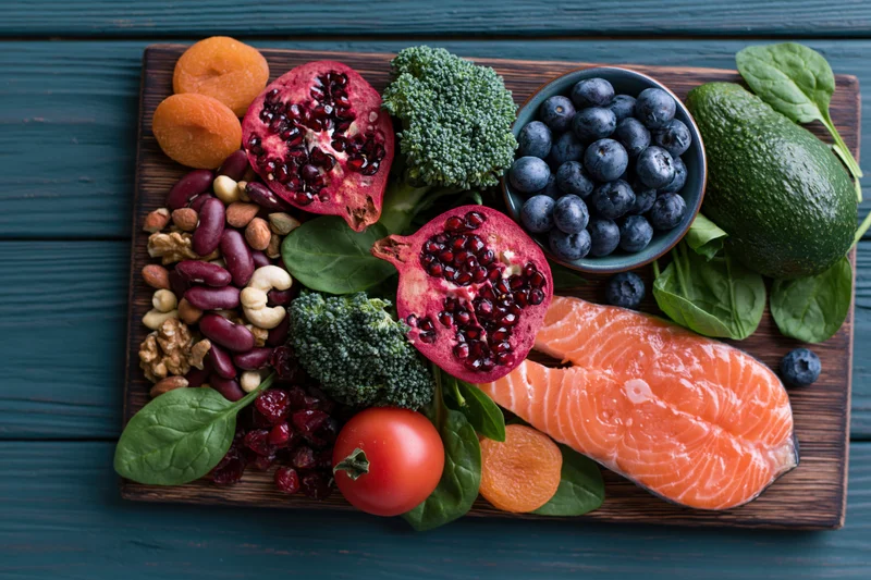 Overhead flat lay of salmon, pomegranate, broccoli, blueberries, avocado, spinach, tomato, apricots, beans, nuts and cranberries arranged on a dark wooden cutting board