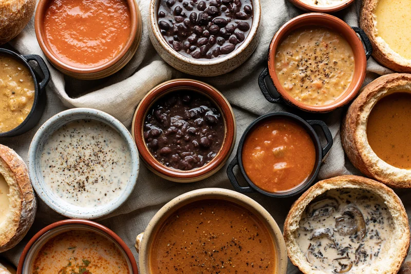 Overhead flat lay of assorted soups and stews in ceramic bowls on a linen cloth