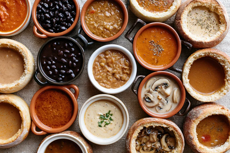 Overhead flat lay of various soups and stews in ceramic bowls and bread bowls on a textured surface