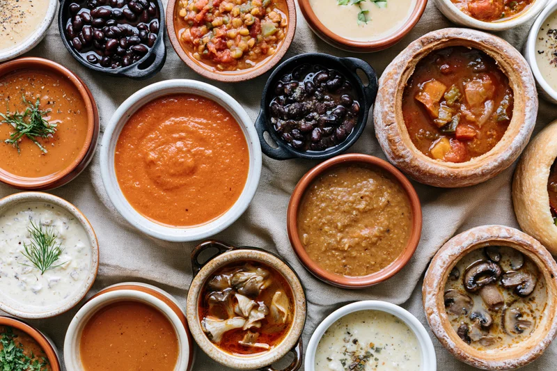 Overhead flat lay of various soups and stews in ceramic bowls on a linen cloth