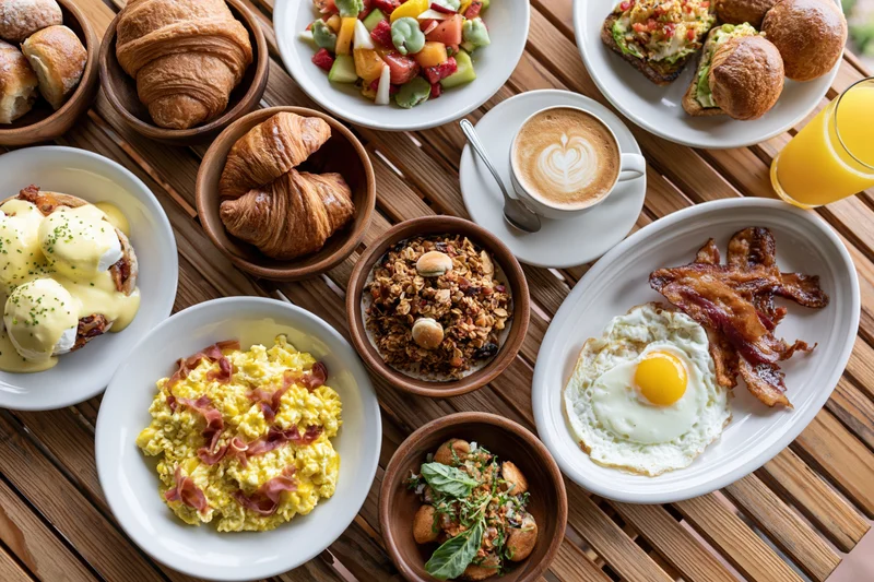 Overhead flat lay of a full brunch spread on a slatted wooden table with croissants, eggs, bacon, avocado toast, granola, fruit salad, coffee and orange juice